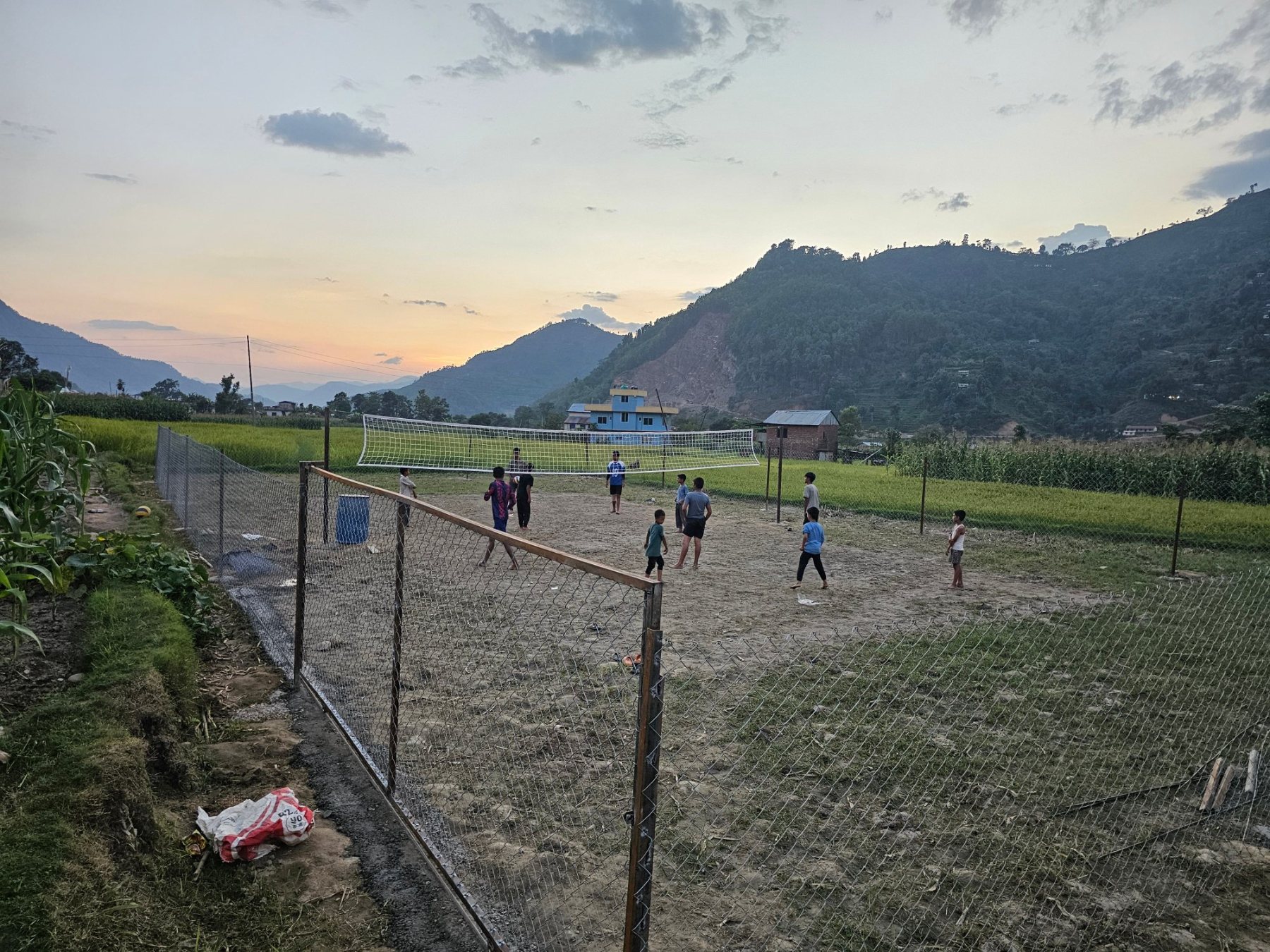 Young people playing volleyball on the completed court at Simara at dusk, maize fields and forested ridges behind